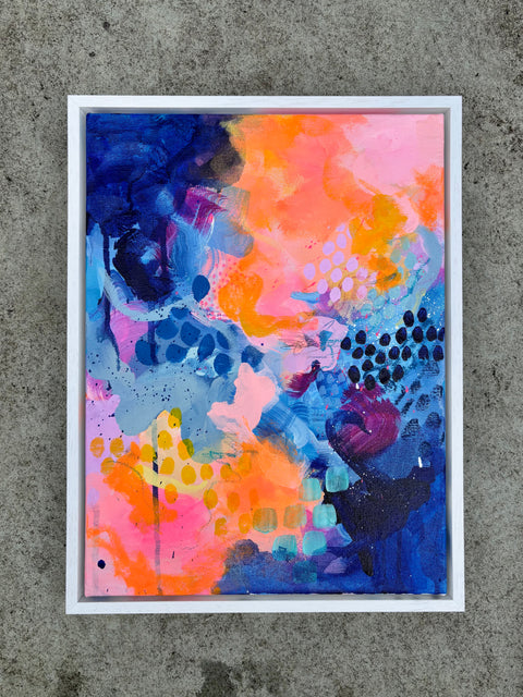 A framed abstract painting with vibrant splashes of blue, pink, orange, and purple, displayed against a textured gray concrete wall.