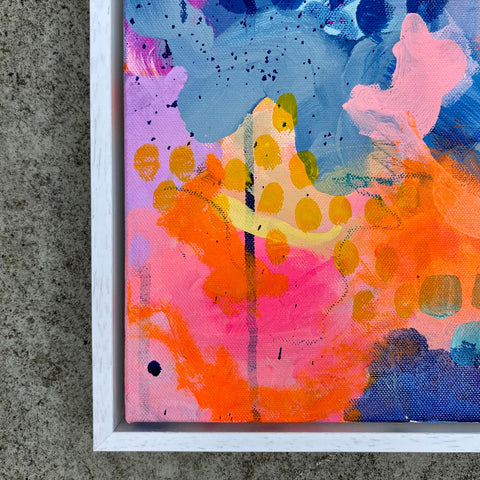 A close-up of a framed abstract canvas painting with bold splashes of pink, orange, blue, purple, and yellow paint, set against a textured gray concrete background.