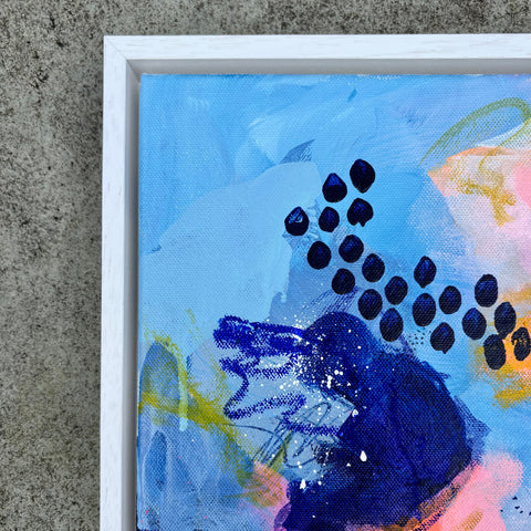 Abstract framed canvas painting with expressive brushstrokes in light blue, navy, pink, yellow, and white, shown with a white frame against a textured gray concrete background.
