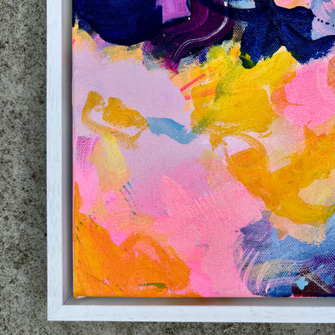 Abstract framed canvas painting with expressive brushstrokes in pink, yellow, blue, purple, and orange, shown in a white frame against a textured gray concrete background.