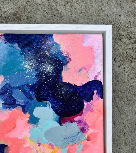 Abstract framed canvas painting with bold brushstrokes in navy, pink, teal, and light blue, shown in a white frame against a concrete surface.