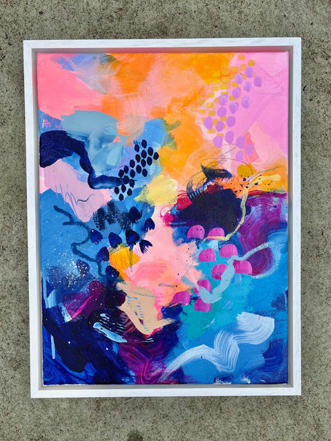 A framed abstract painting featuring a vibrant mix of pink, orange, blue, purple, and black brushstrokes, presented in a white frame against a textured gray concrete wall.
