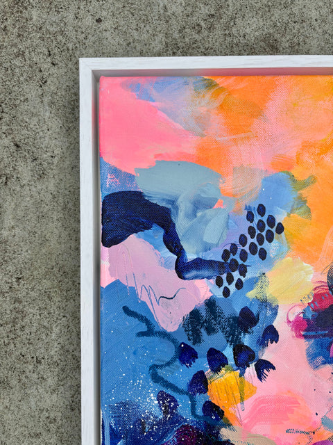 Abstract framed painting with vibrant pink, orange, blue, and purple brushstrokes, shown in a white frame against a textured gray concrete wall.