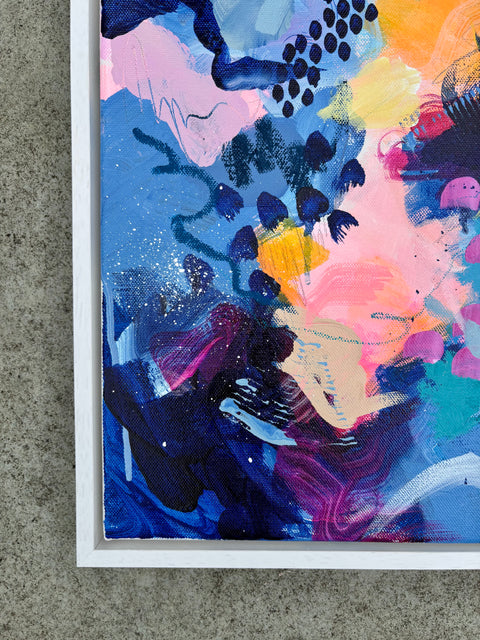 A close-up of a framed abstract painting with expressive brushstrokes in blue, pink, orange, purple, and black, mounted against a textured gray wall.